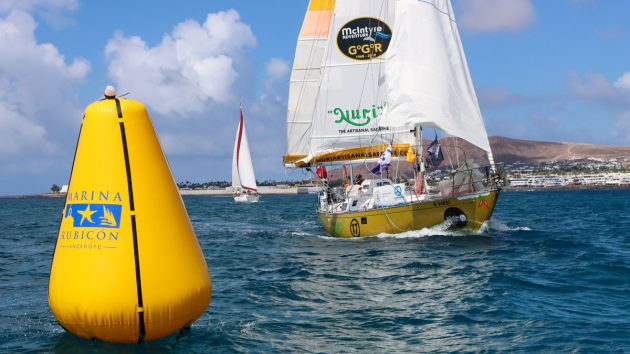 A boat with a yellow hull and white sails, passing by a buoy