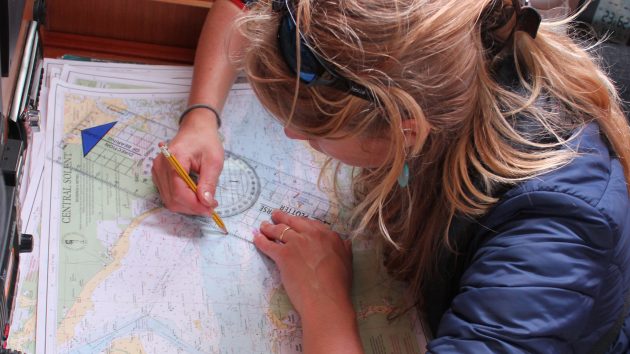 A woman with blonde hair charting a course on a paper chart on a boat