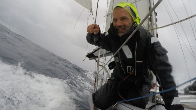 A male sailor in wet weather gear with a yellow hood on the edge of a yacht while sailing through white water