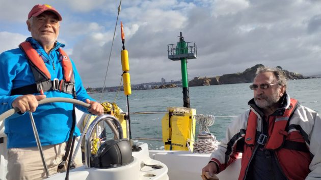 A man wearing a blue t-shirt, red lifejacket and a red and yellow cap at the wheel of a yacht