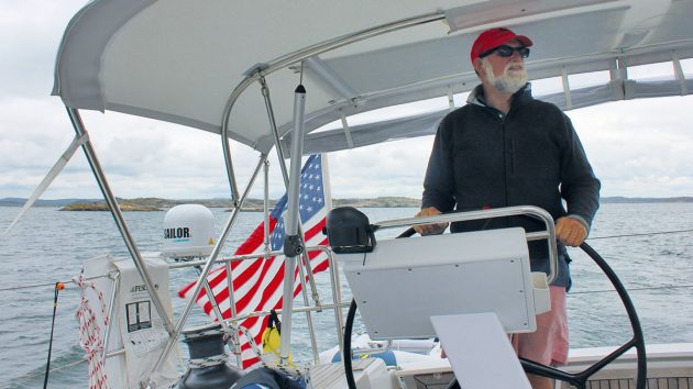 A man wearing a red baseball cap and black glasses sailing a boat with a satellite antenna on the railing for marine satellite communication
