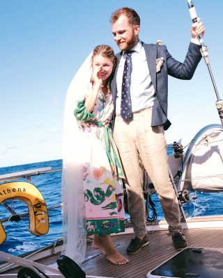Philip and Evelina are grinning and standing on deck. Evelina is wearing a white veil and wiping away a tear. The sky is blue and the couple are wearing formal clothes.