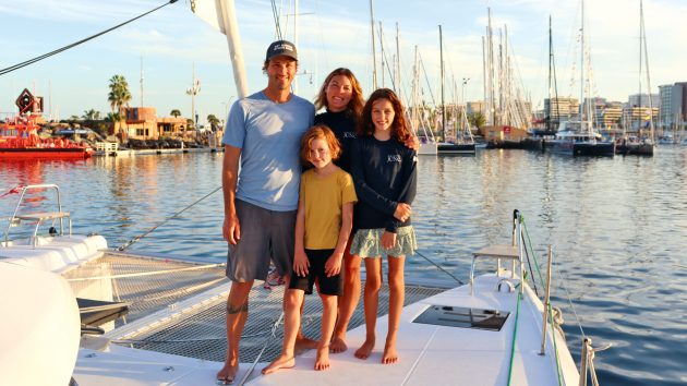The Hunt family is standing on their yacht, smiling. There are palm trees and gentle waters behind them.