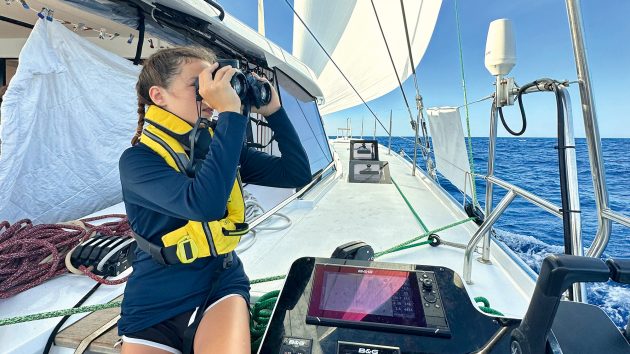 Sonnen sitting on the yacht, wearing a lifejacket and looking through binoculars at the horizon. It is fair weather.
