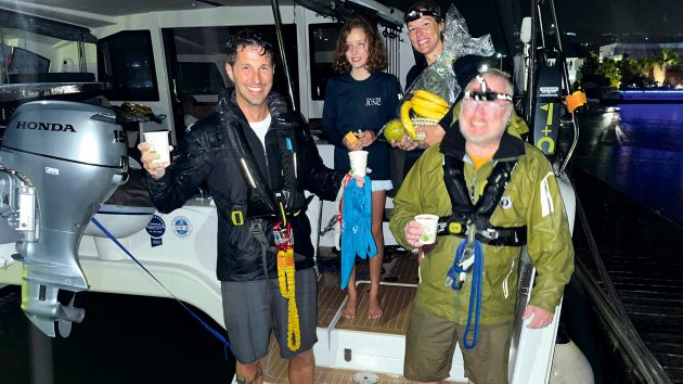 The family are smiling with Max aboard their catamaran. It's night, and they are wearing waterproof clothing.
